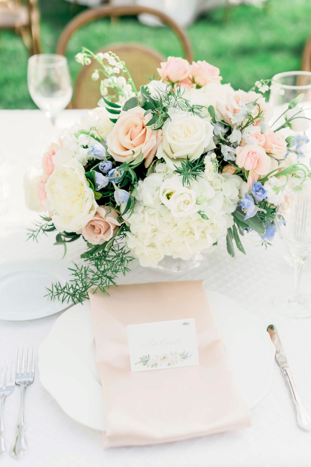 Elegant floral centerpiece with blush roses and white hydrangeas on a formal place setting with pink napkin.