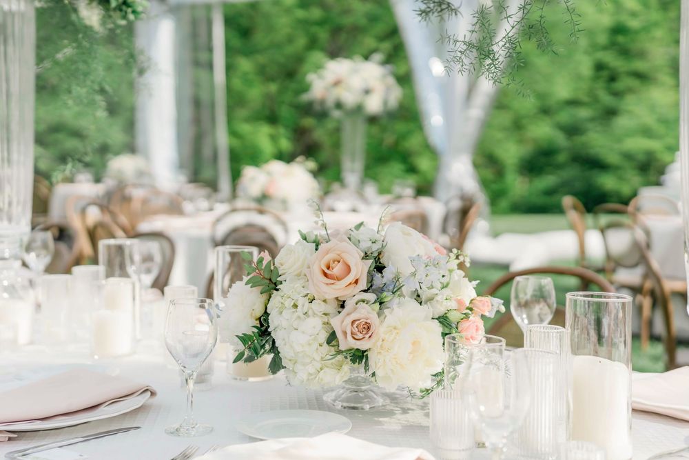 An elegant table setting features a delicate centerpiece of blush roses and white flowers surrounded by glassware.