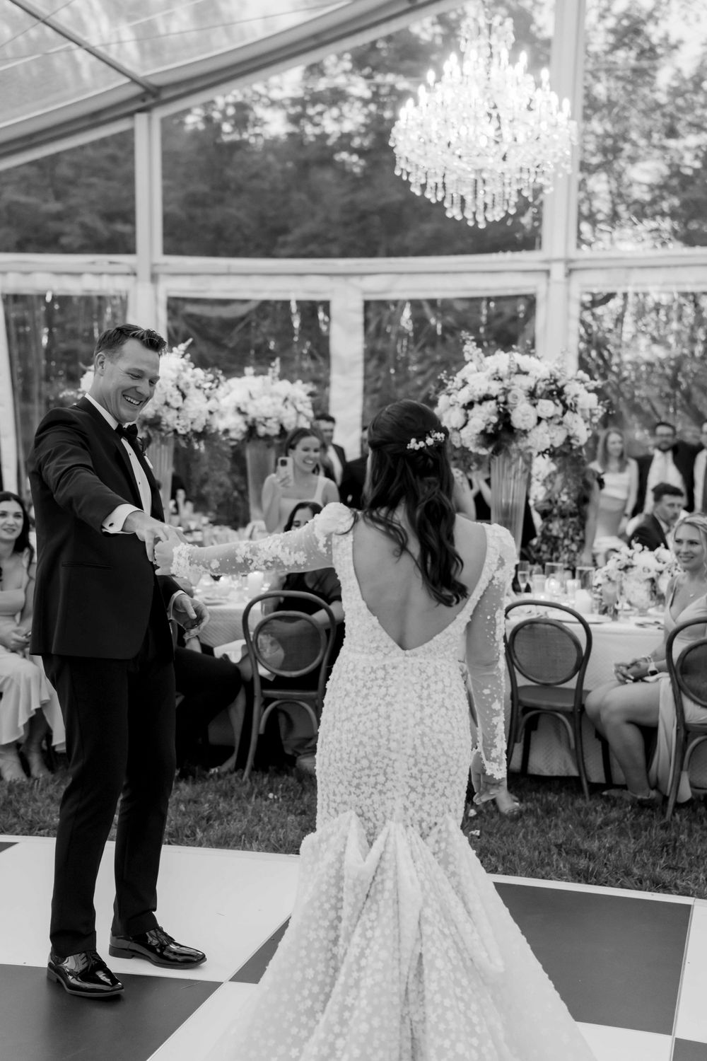 A couple shares their first dance at a wedding reception under an elegant crystal chandelier in a clear tent pavilion.
