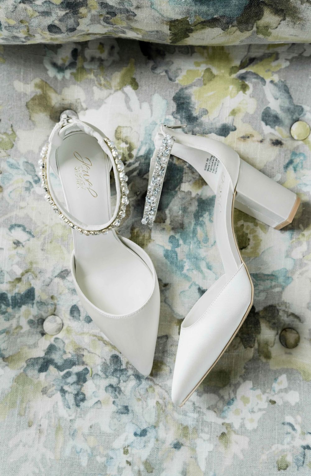 White bridal heels with pearl ankle straps photographed on a watercolor-style background.