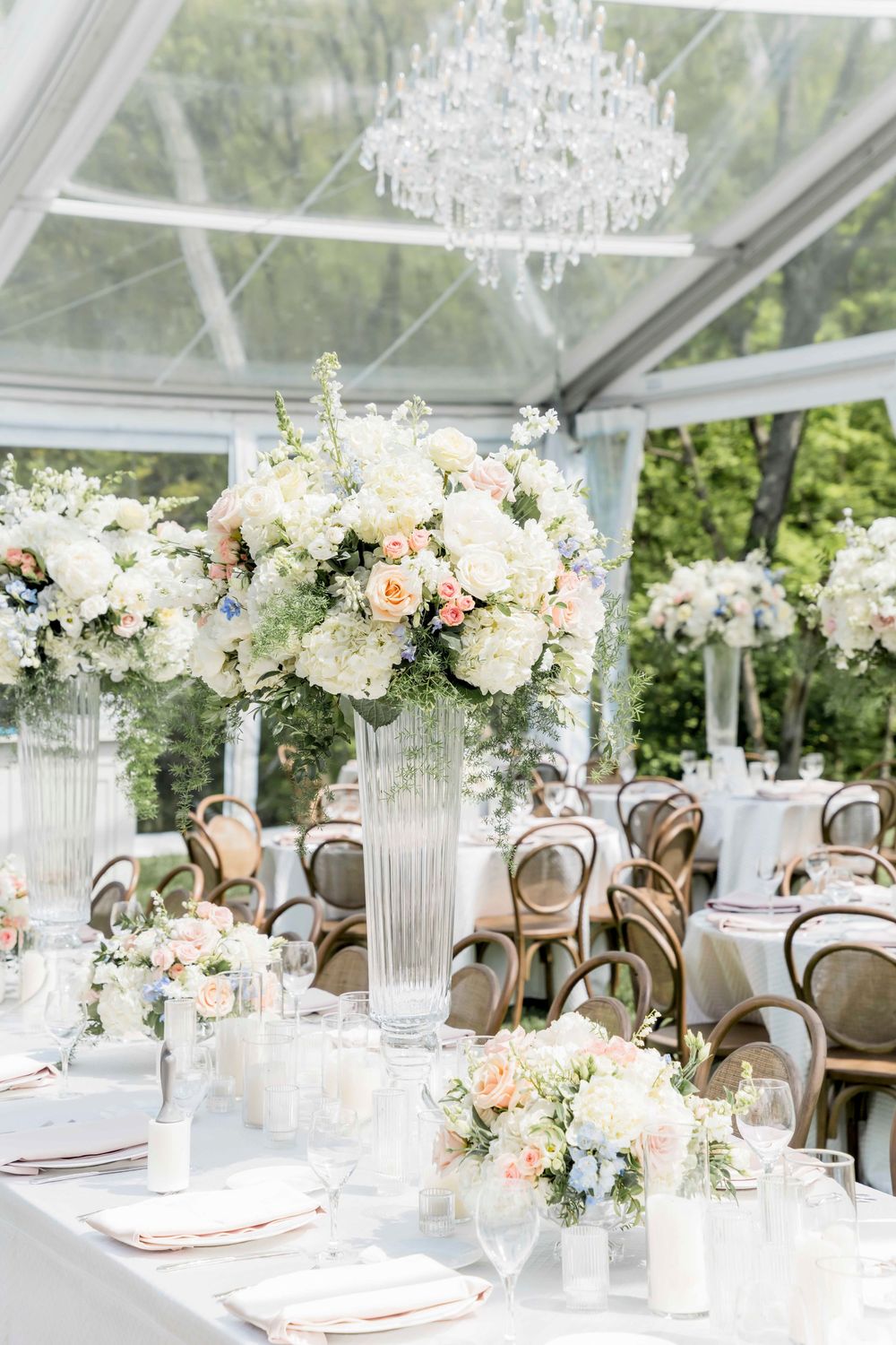 Romantic tented wedding reception with crystal chandelier and tall floral centerpieces in a clear-top marquee.