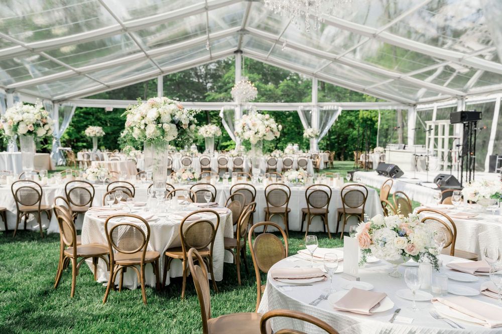 Outdoor wedding reception under a transparent tent with white floral decor and rustic wooden chairs.
