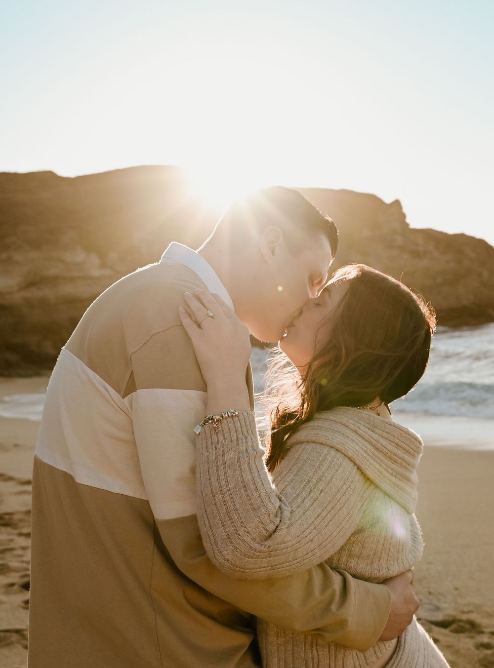 Romantic couple in cozy sweaters sharing a tender moment on the beach at sunset with golden light streaming behind them, in Pacifica,CA