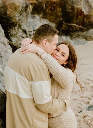 A couple in neutral sweaters sharing an intimate embrace on a rocky beach at sunset.
