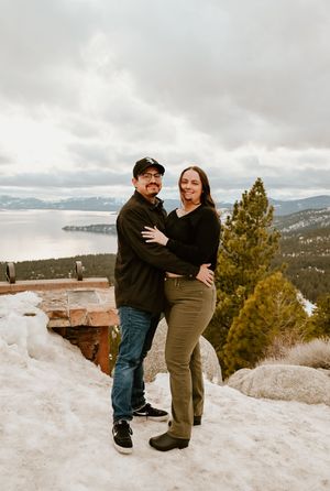 A couple embraces on a snowy mountain overlook with Lake Tahoe and evergreen trees visible in the background, in North Tahoe