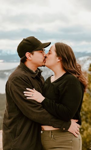 A couple in casual winter clothing share a romantic kiss on a scenic mountain overlook with cloudy skies in the background, in Tahoe