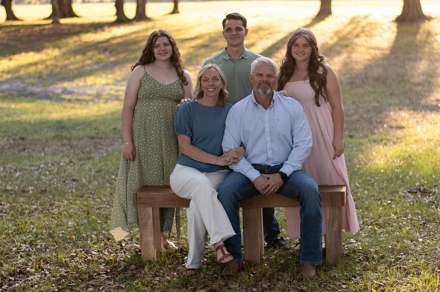 Four people sit closely together on green grass surrounded by trees in a casual outdoor setting.