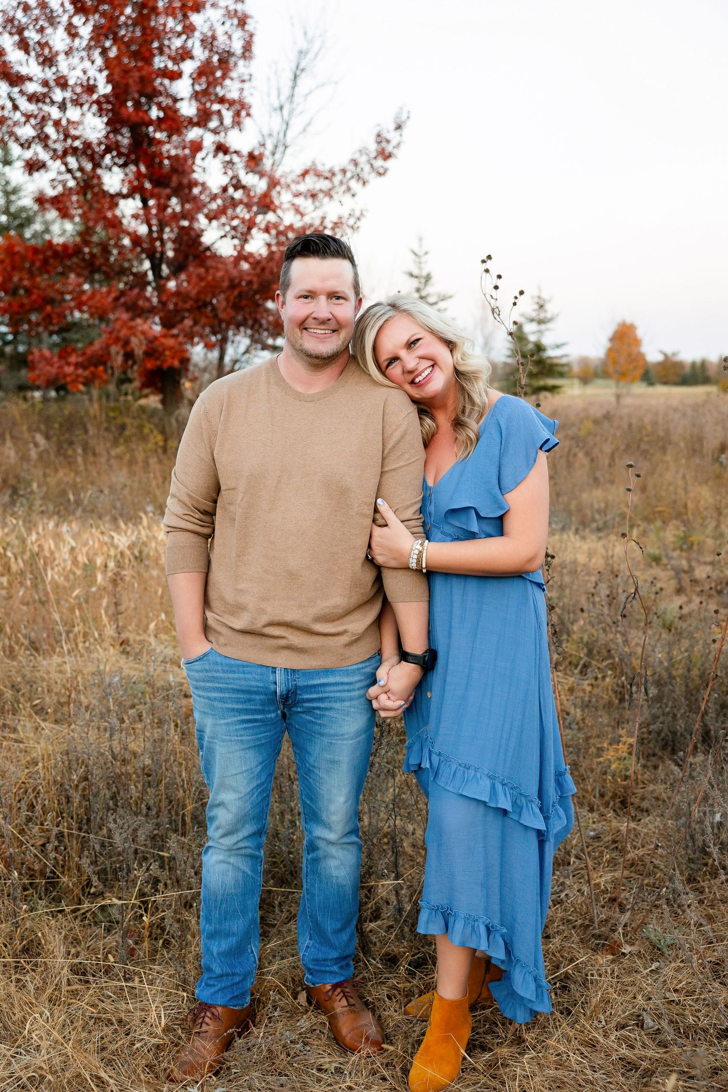 A couple poses together in a field during autumn with red foliage and golden grass in the background.