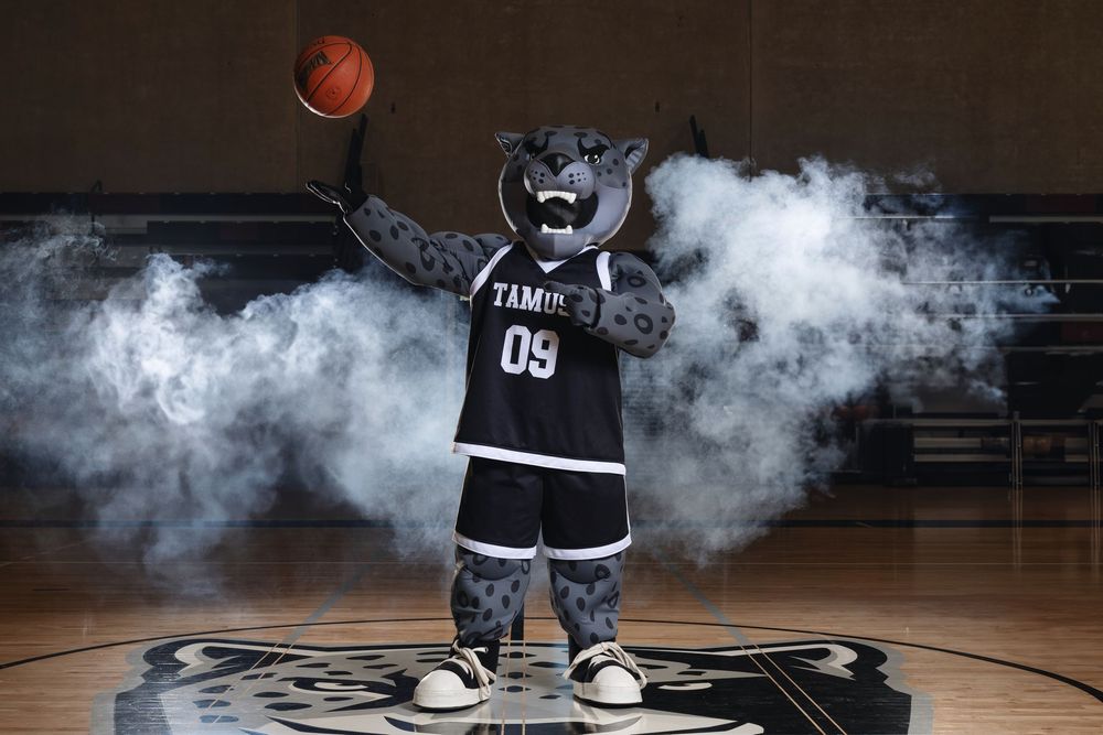 A costumed basketball team mascot in black and white uniform poses with a basketball on court surrounded by smoke effects.