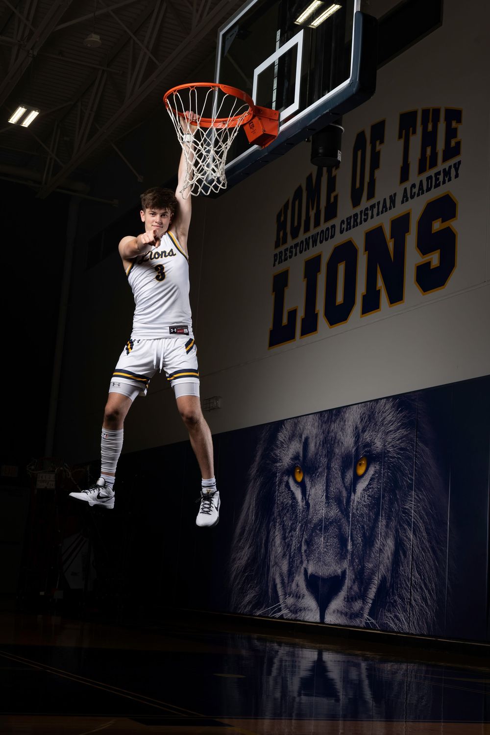 Basketball player leaps high for a slam dunk in front of a Lions team logo wall at a Christian academy gymnasium.