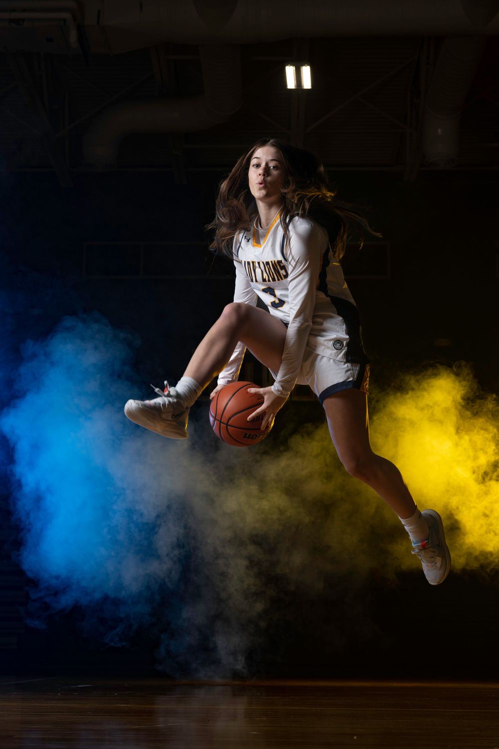 Basketball player jumps with ball against dramatic blue and yellow smoke effects in a dark gymnasium.