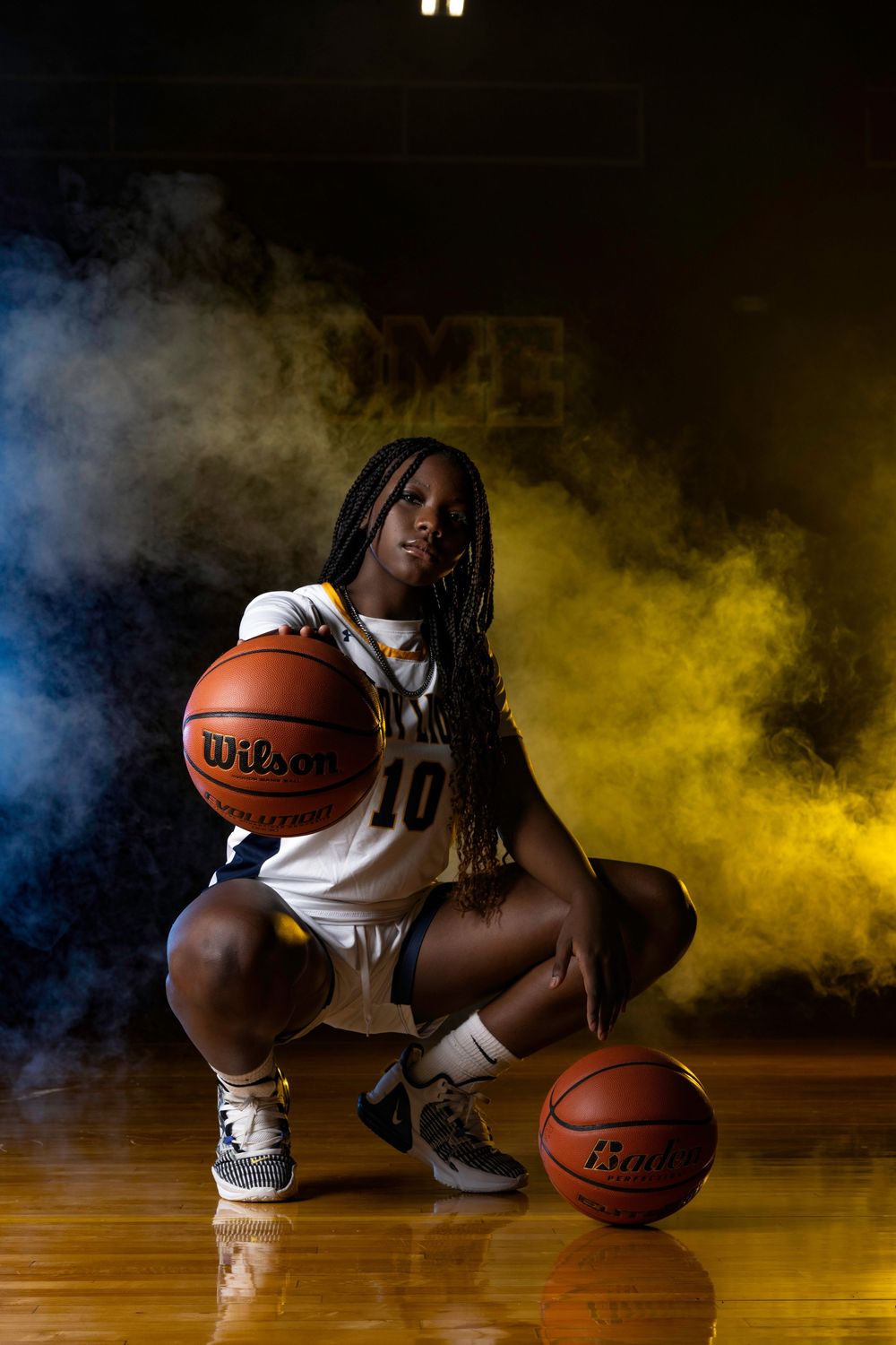 Basketball player kneels with two Wilson basketballs on wooden court surrounded by dramatic yellow smoke effect.