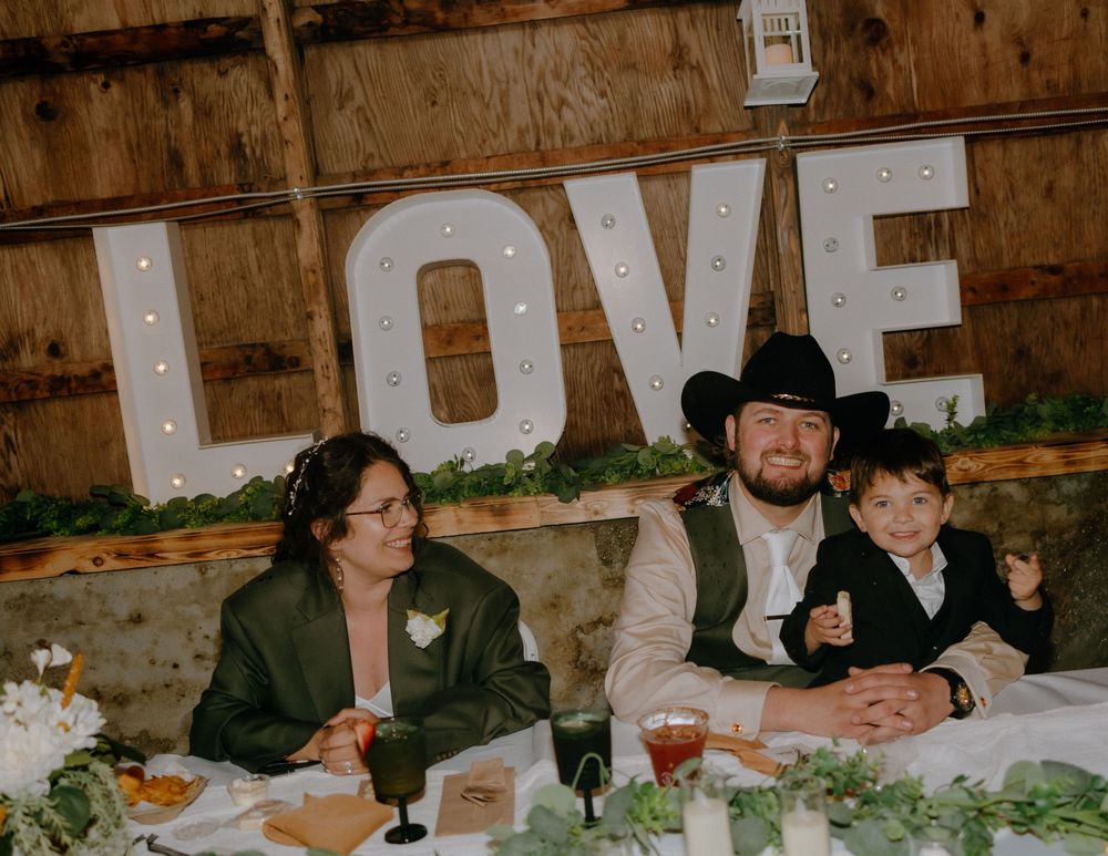 Family sits at a rustic reception table with wooden LOVE sign and floral decorations behind them at a wedding celebration.