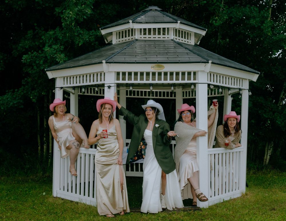A group poses together in white dresses and sun hats while standing around an elegant white outdoor gazebo in a garden setting.