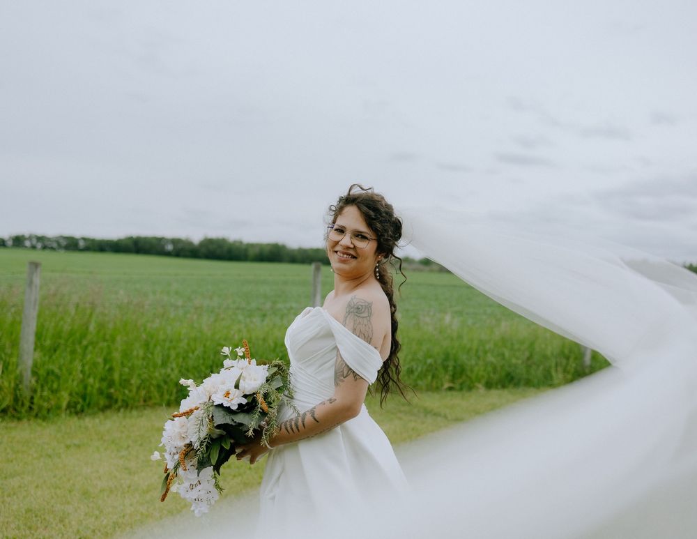 A joyful bride in a white off-shoulder wedding gown holds a floral bouquet against a green rural landscape as her veil flows.