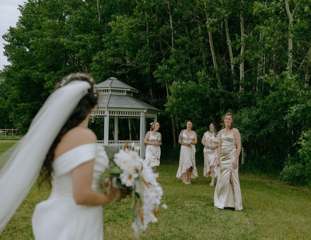 A wedding party gathers near a gazebo surrounded by trees with the bride in a white dress holding flowers.