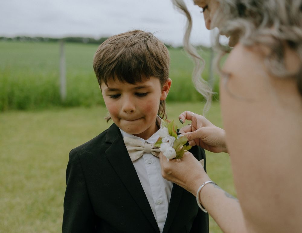 A young boy in a black suit with a bowtie smiles as someone adjusts his boutonniere outdoors.