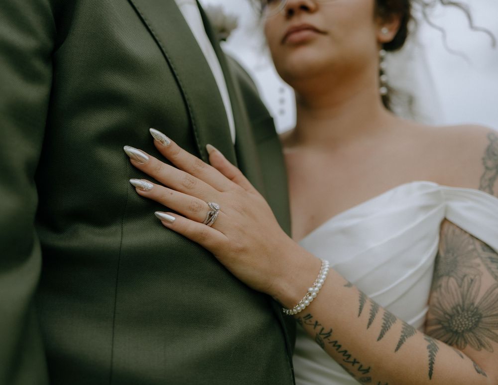 Close-up of a bride's tattooed arm and wedding ring against a green suit jacket.