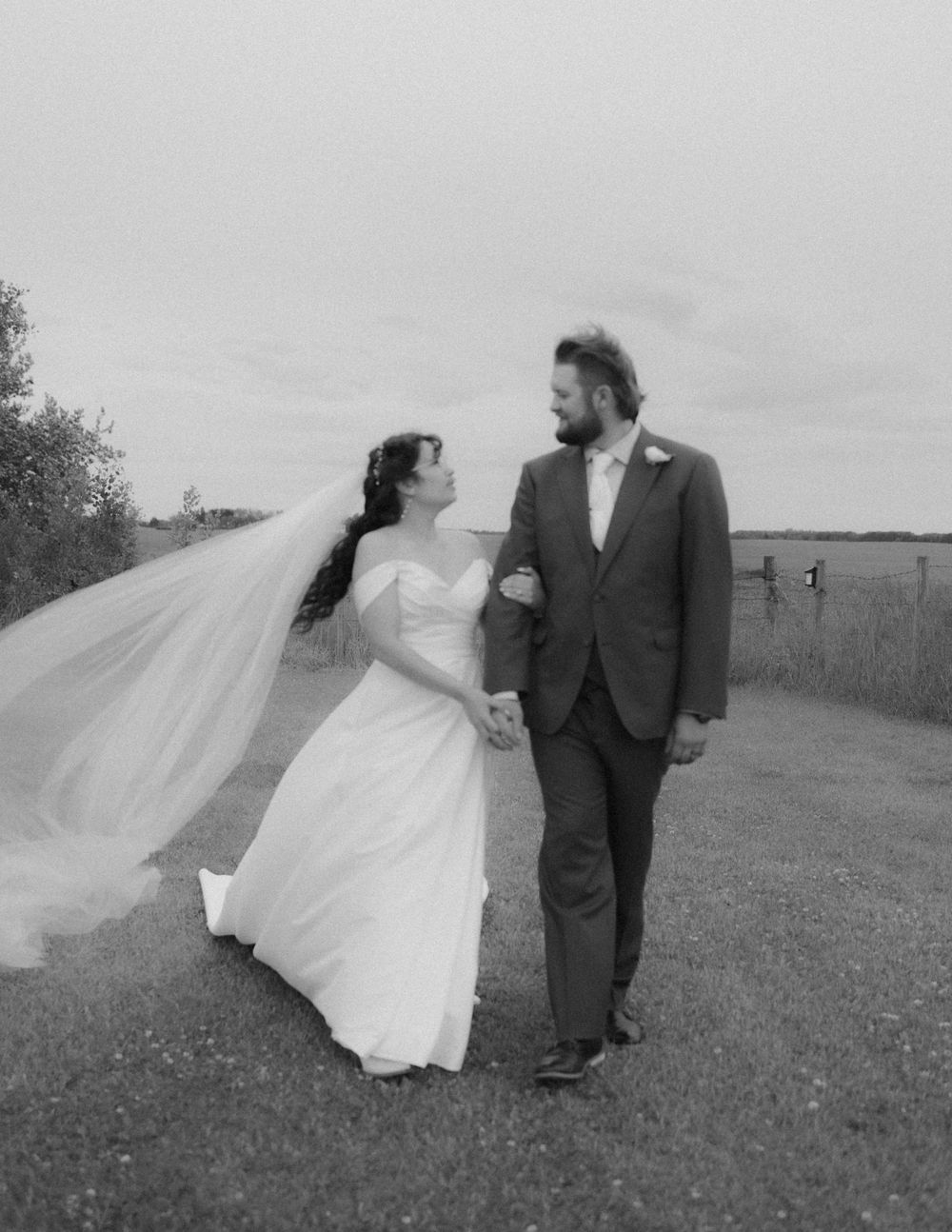 A bride in a flowing white dress and groom in a dark suit walk together on a grassy path under an overcast sky.