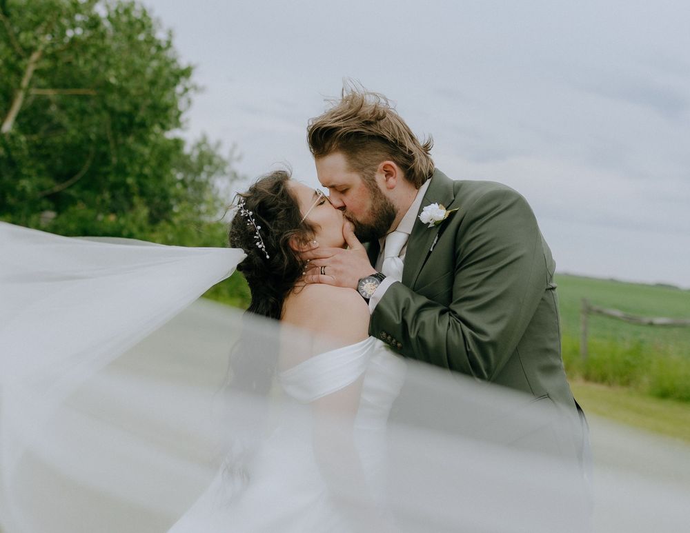 Couple embraces in romantic kiss outdoors with white veil flowing in the wind against a green rural landscape and overcast sky.