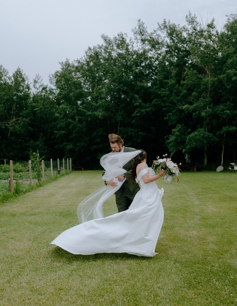 A couple in wedding attire dance joyfully on a grassy lawn with a forest backdrop as the bride's dress flows in the wind.
