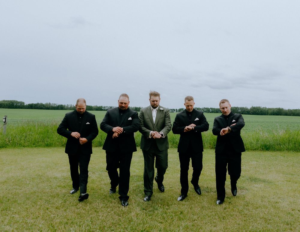 A group of five men in matching black suits stand together in a grassy field on a cloudy day.