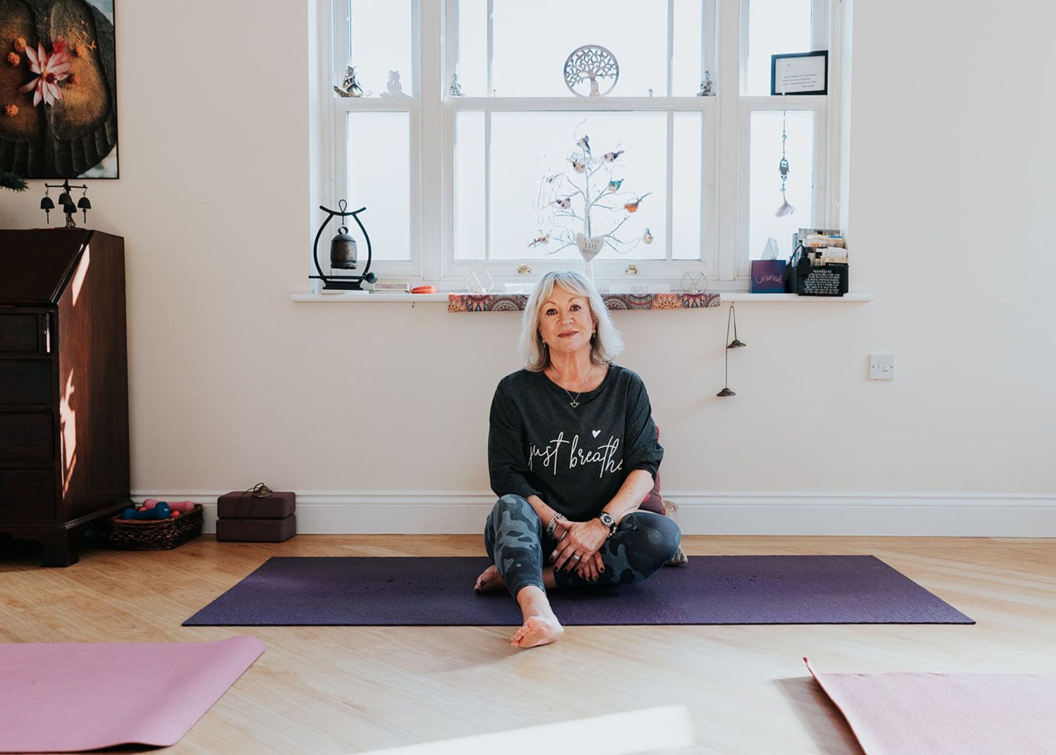 Someone sits cross-legged on a purple yoga mat in a bright minimalist room with decorative window sill items.