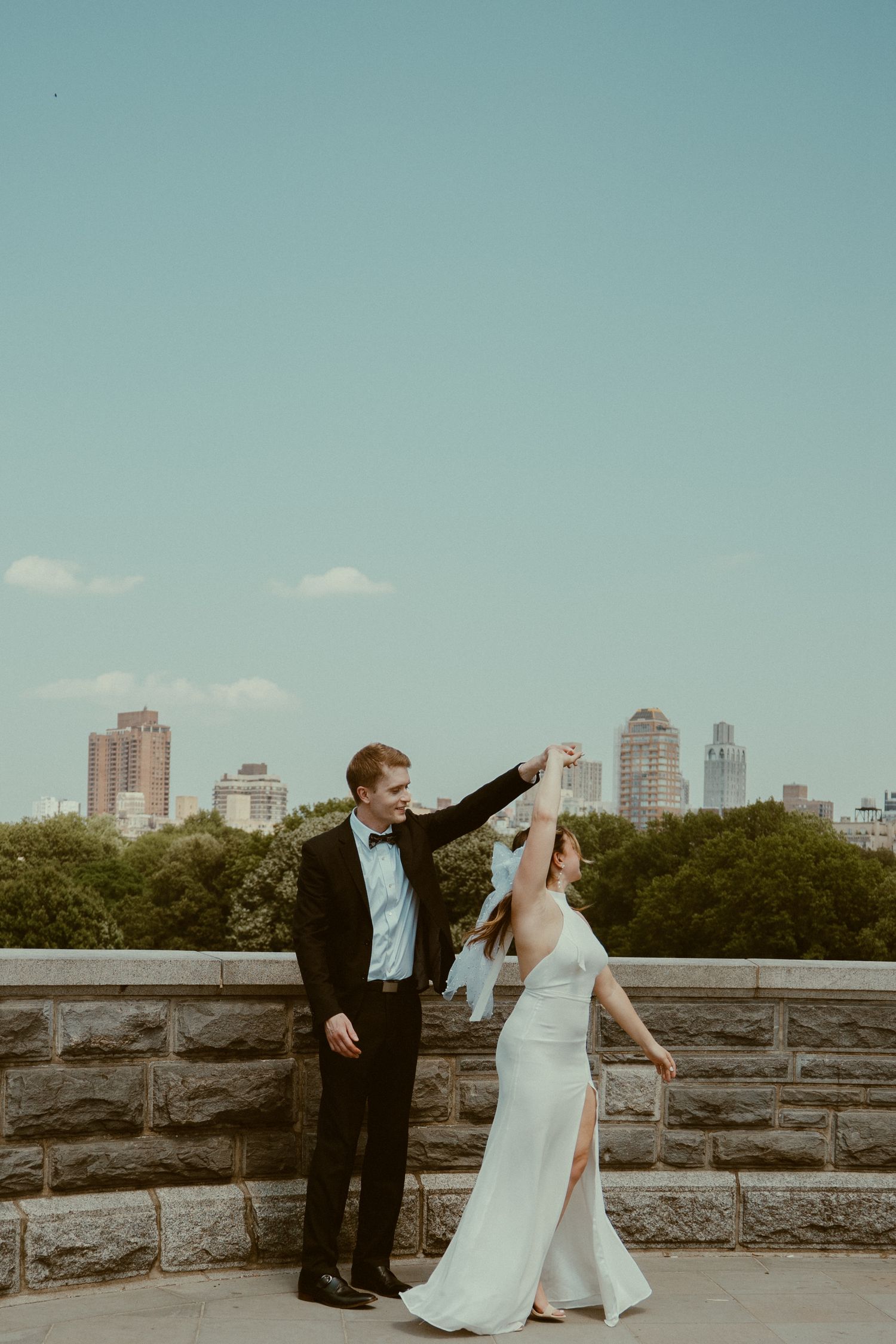 Couple in wedding attire walking along stone wall with city skyline in background across four sequential photos.
