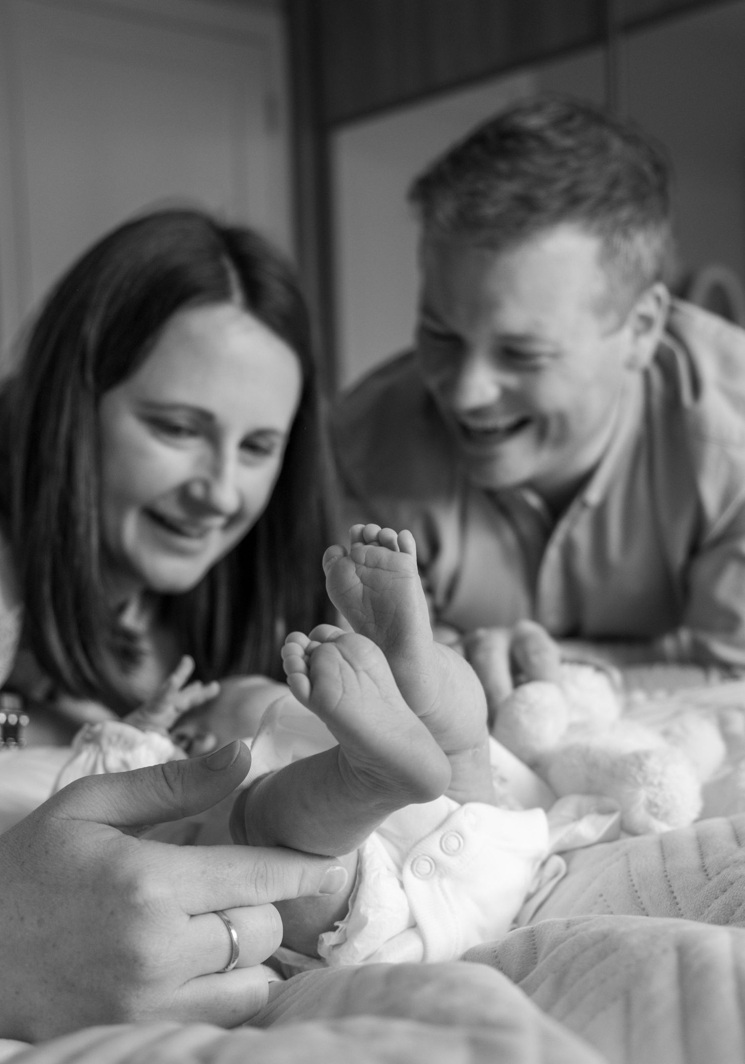 Black and white photo of smiling parents admiring their newborn baby's tiny feet on a bed.
