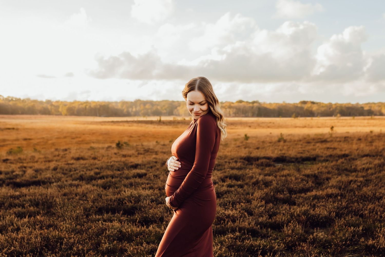 A pregnant woman standing alone in the New Forest, holding her belly and smiling.