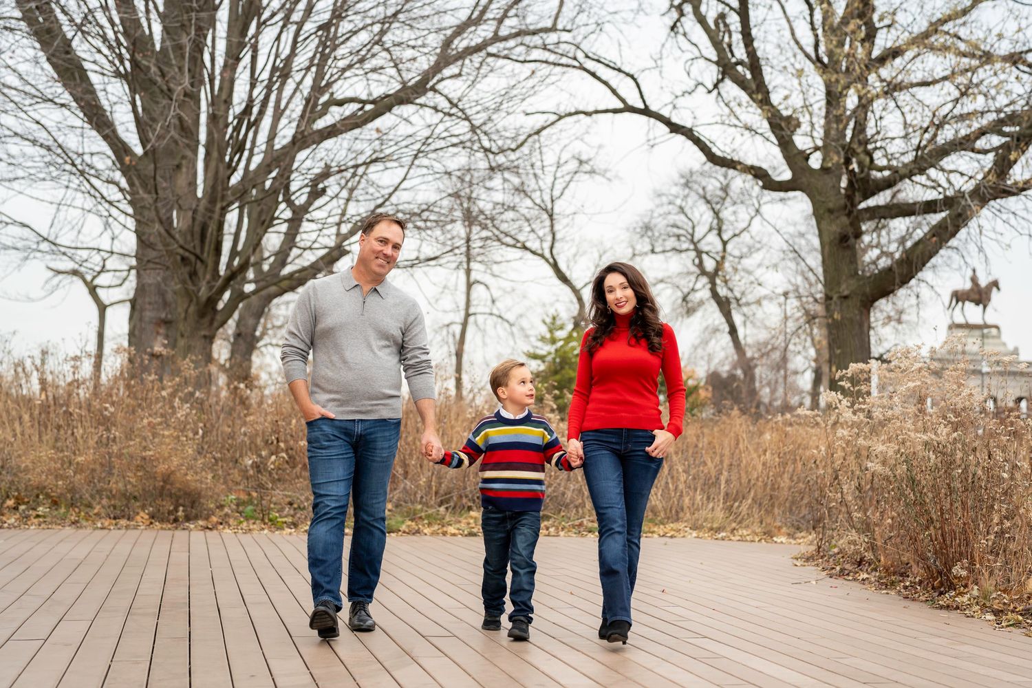Family holding hands while walking on wooden boardwalk through autumn nature trail in park.