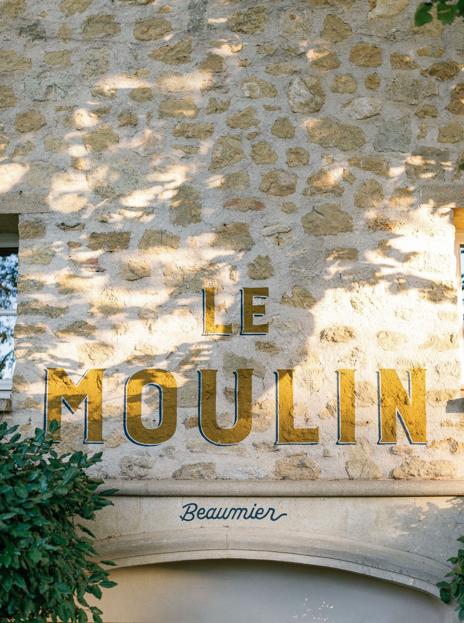 Vintage 'Le Moulin' text painted on a rustic stone wall with dappled sunlight and green foliage below.