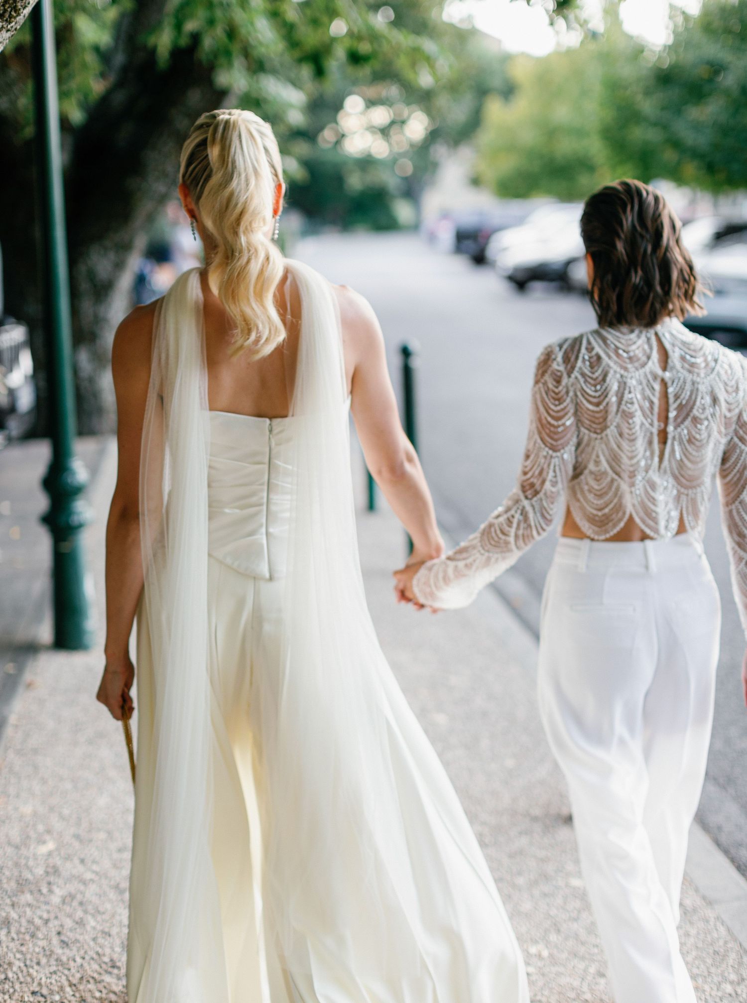 A group of people in white wedding dresses walk down a tree-lined path during a bridal photoshoot.