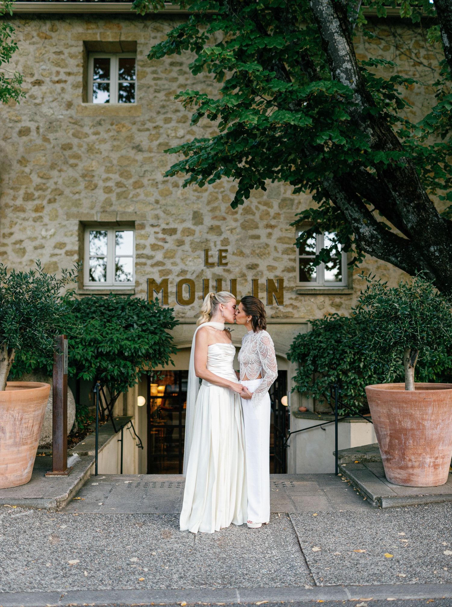 Two people in white dresses share an intimate moment outside a rustic stone building at dusk.