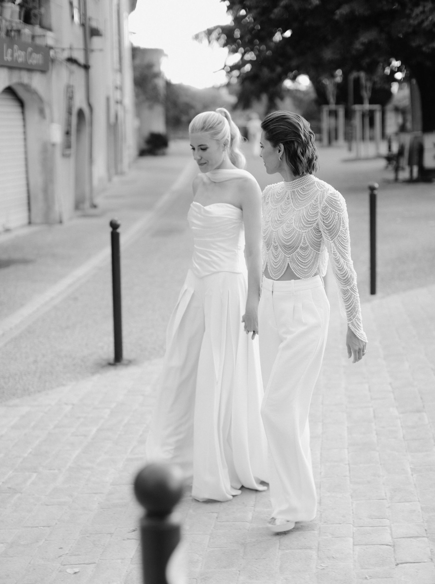 Two people in white dresses walk together down a cobblestone street in black and white photography.