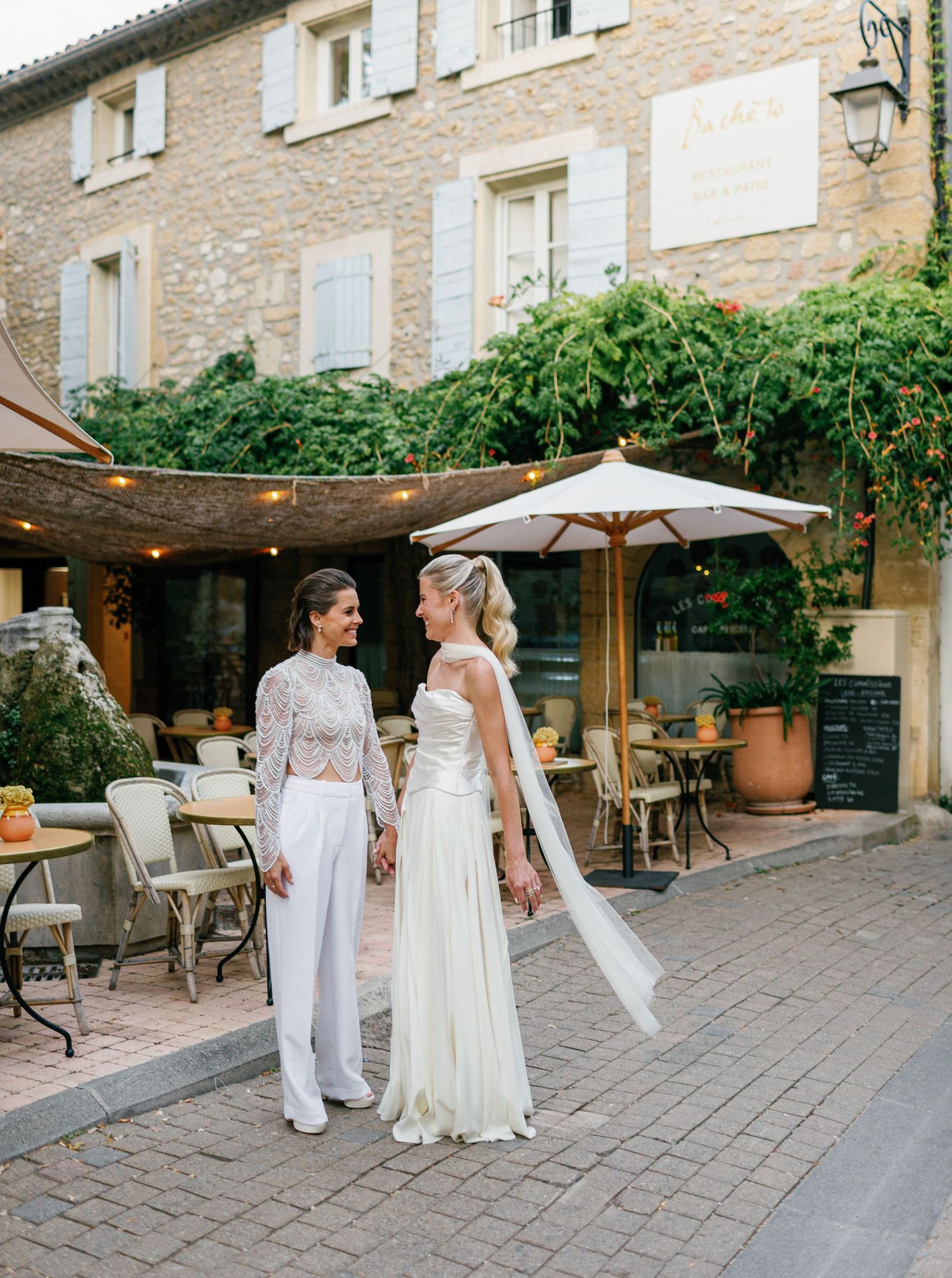 A group in white attire gathers on a stone patio with string lights and outdoor furniture at a wedding venue.