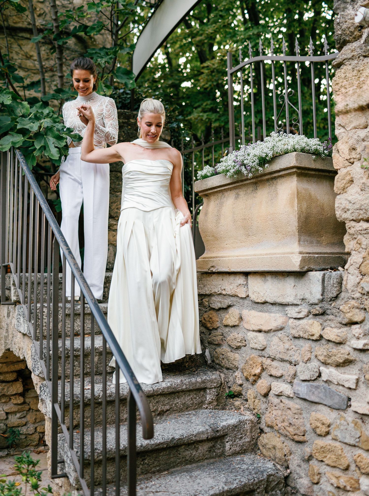 A couple in wedding attire descends stone steps in a garden setting with lush greenery and an iron railing.