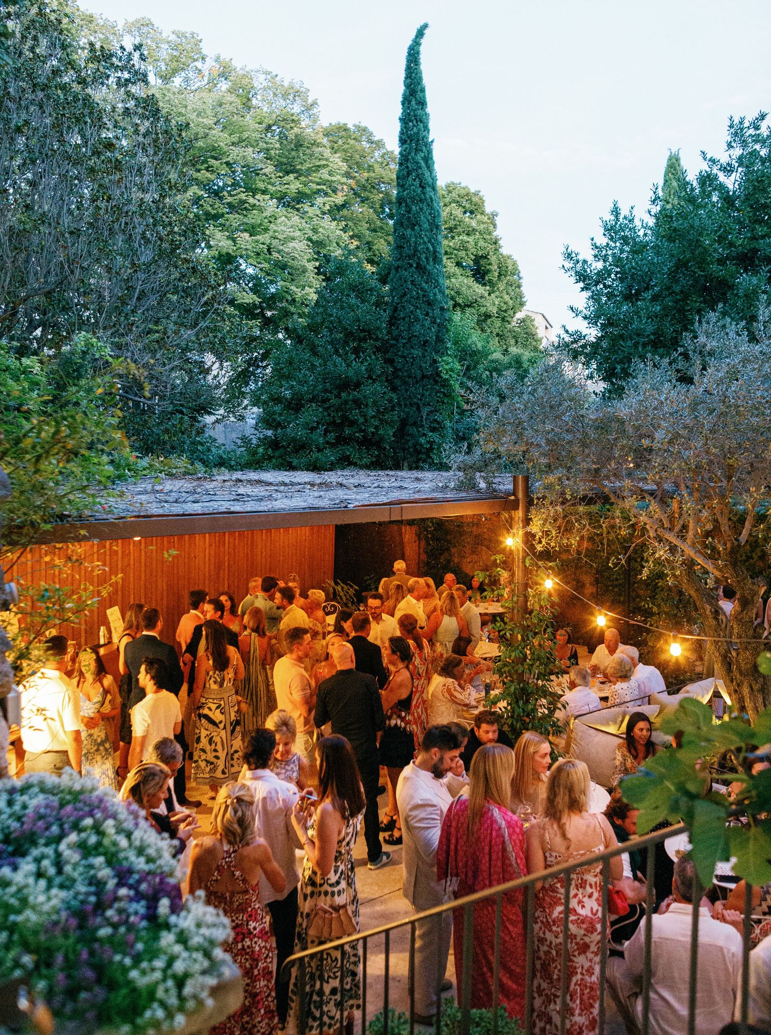 Guests gather at an outdoor evening garden party with string lights and cypress trees creating an enchanting atmosphere.
