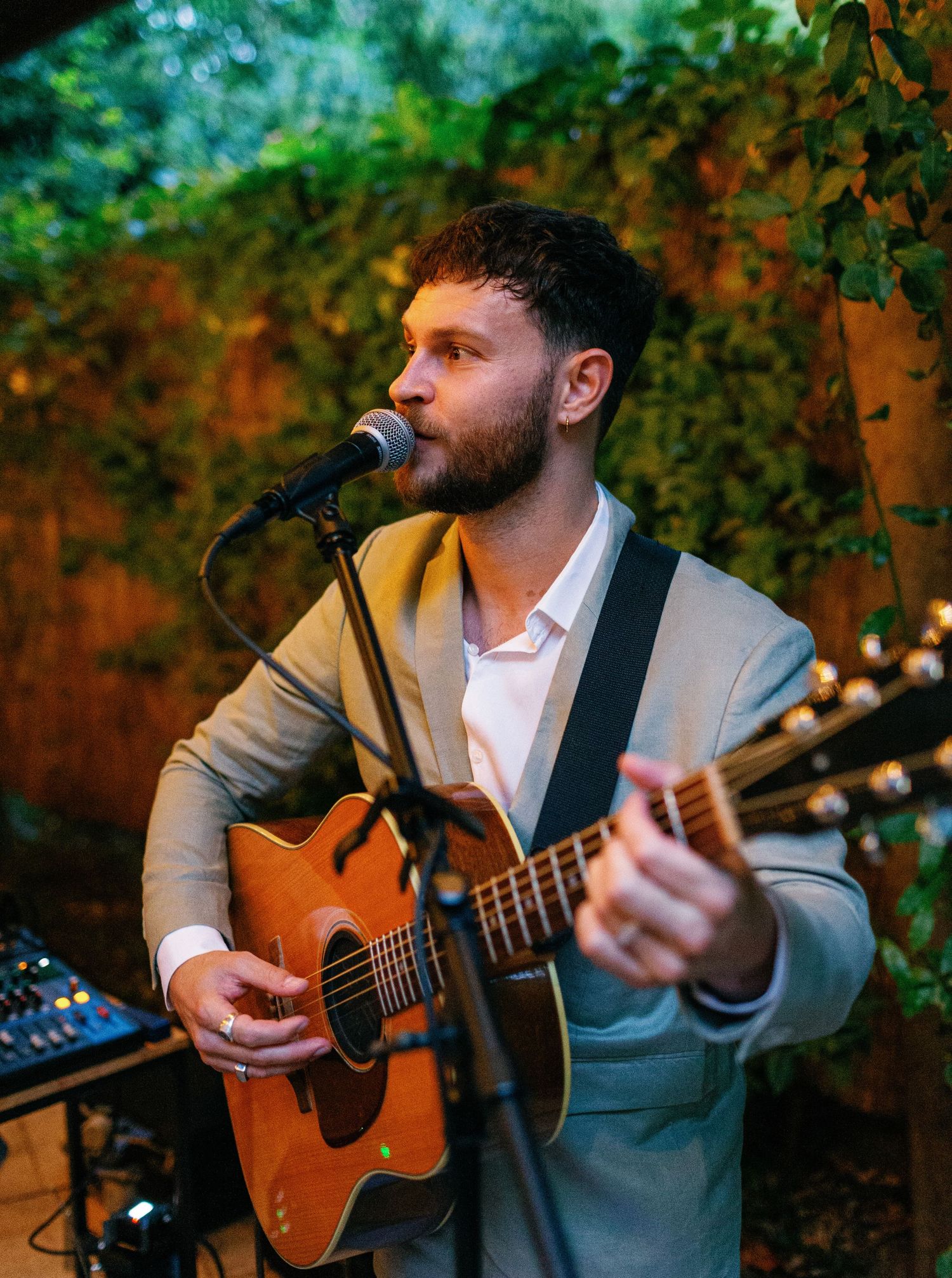 A musician in a gray suit performs with an acoustic guitar in a lush outdoor garden setting with warm lighting.