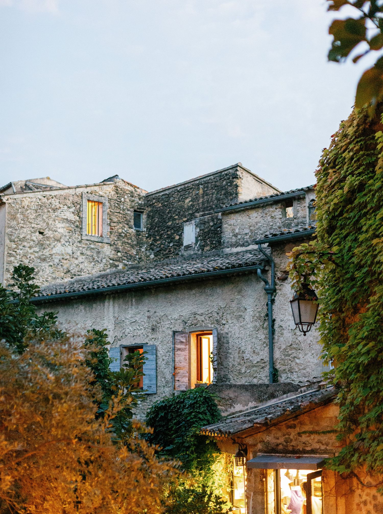 Rustic stone house with glowing windows and ivy-covered walls at dusk in a charming European village setting.