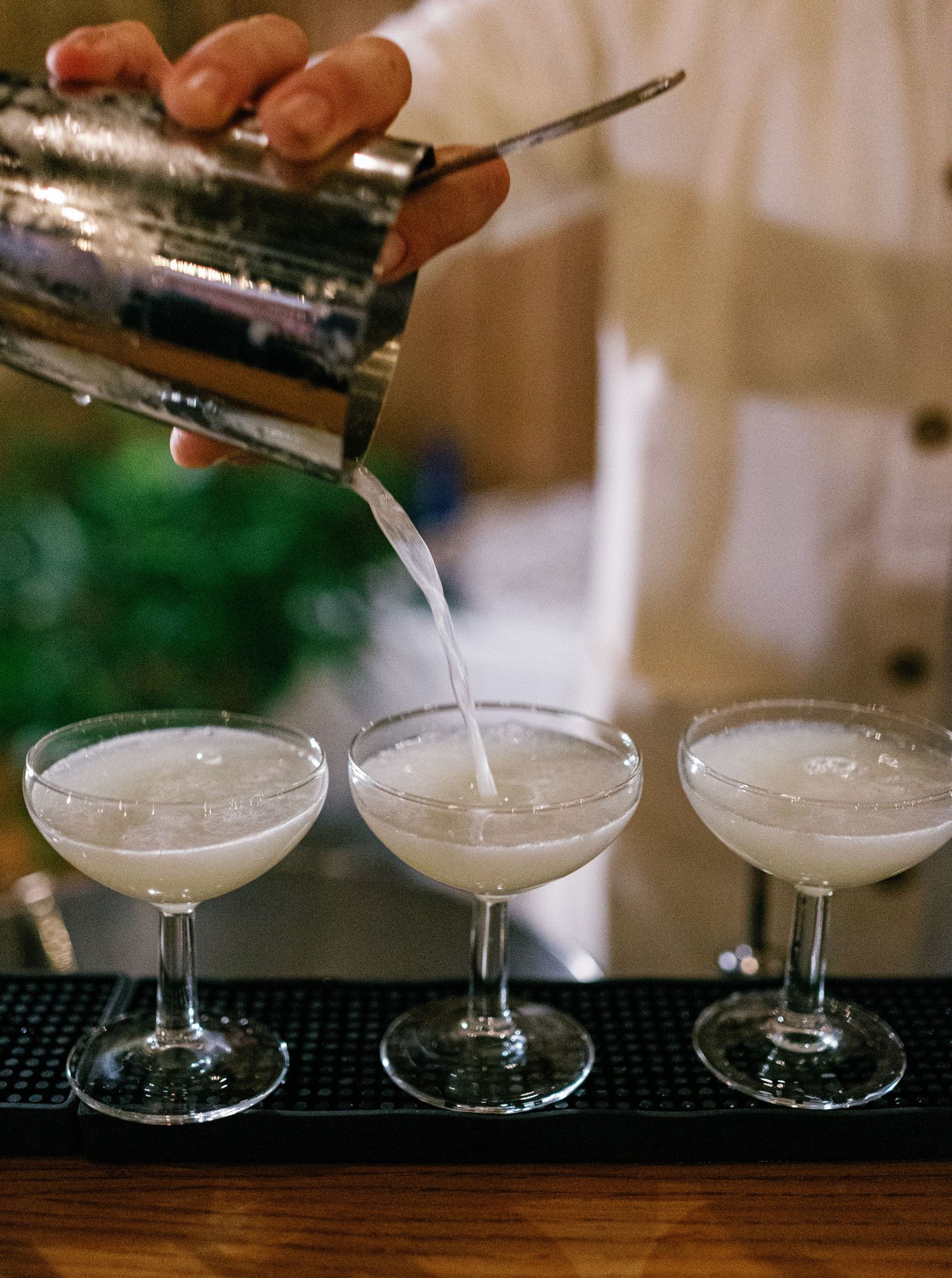 A cocktail shaker pours white frothy drinks into three coupe glasses lined up on a bar mat.