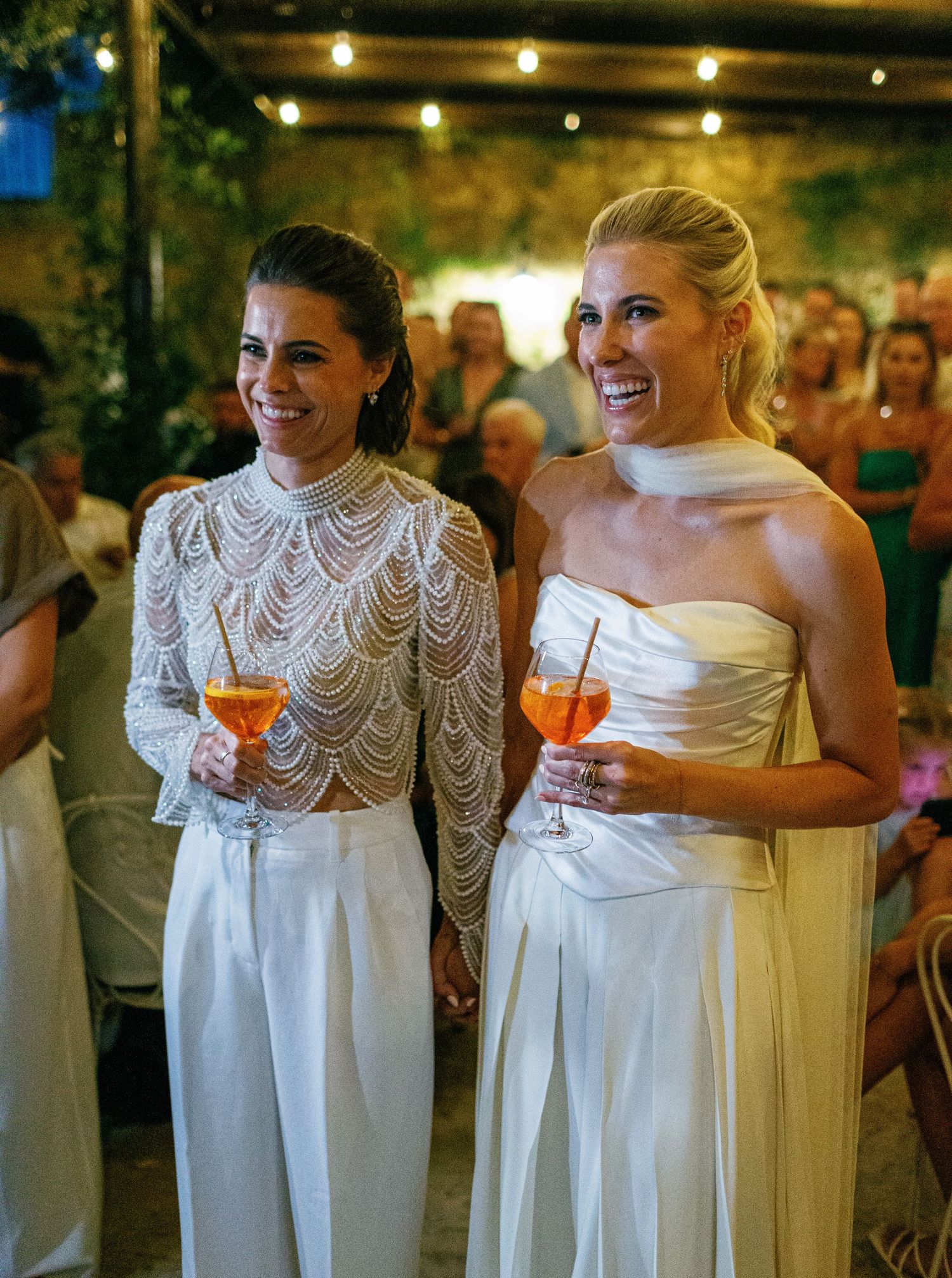 Bridesmaids in white dresses with lace details share laughter and drinks during an evening wedding reception.