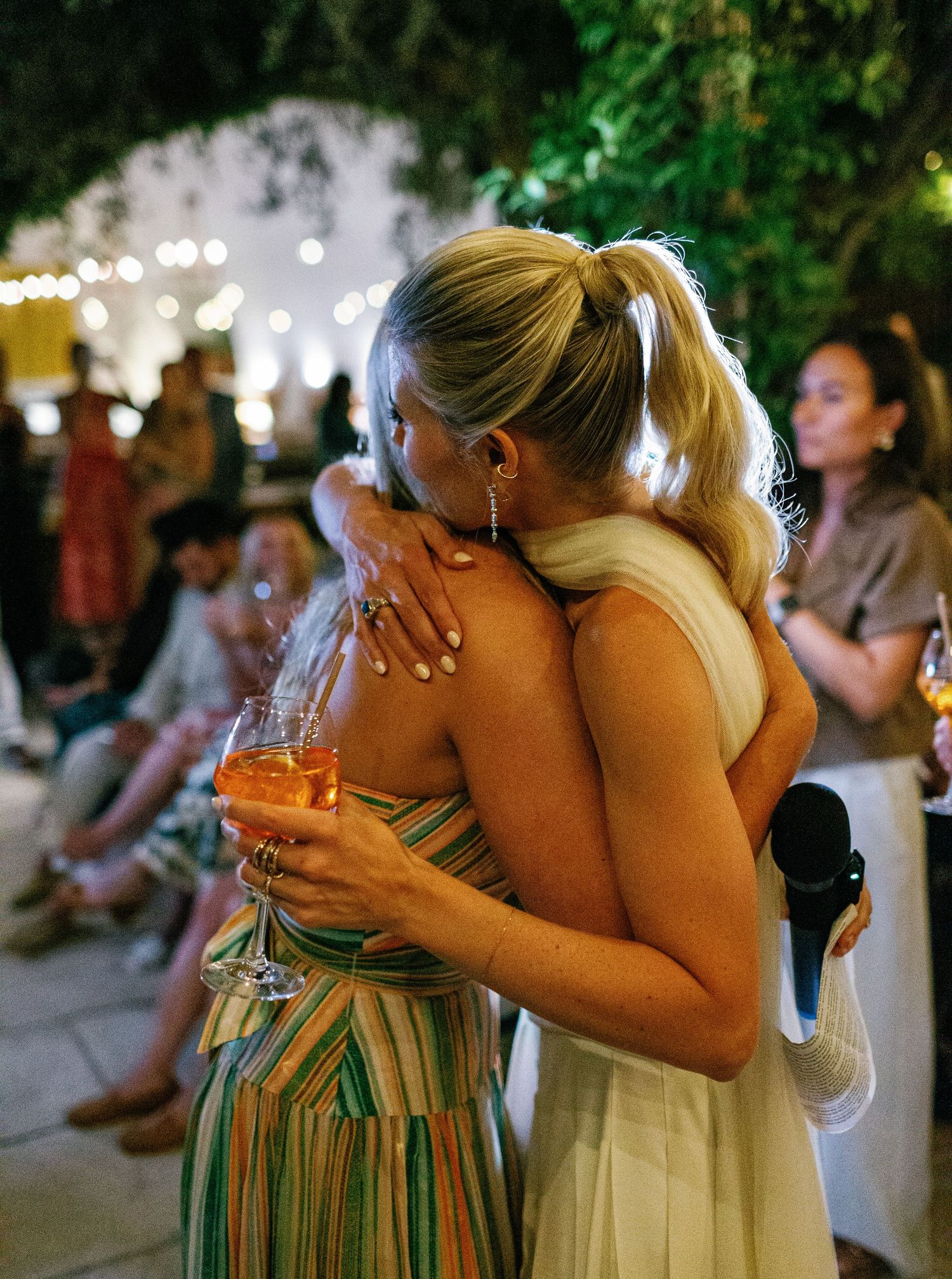 Two people share an emotional embrace at an outdoor evening event with twinkling lights and greenery in the background.