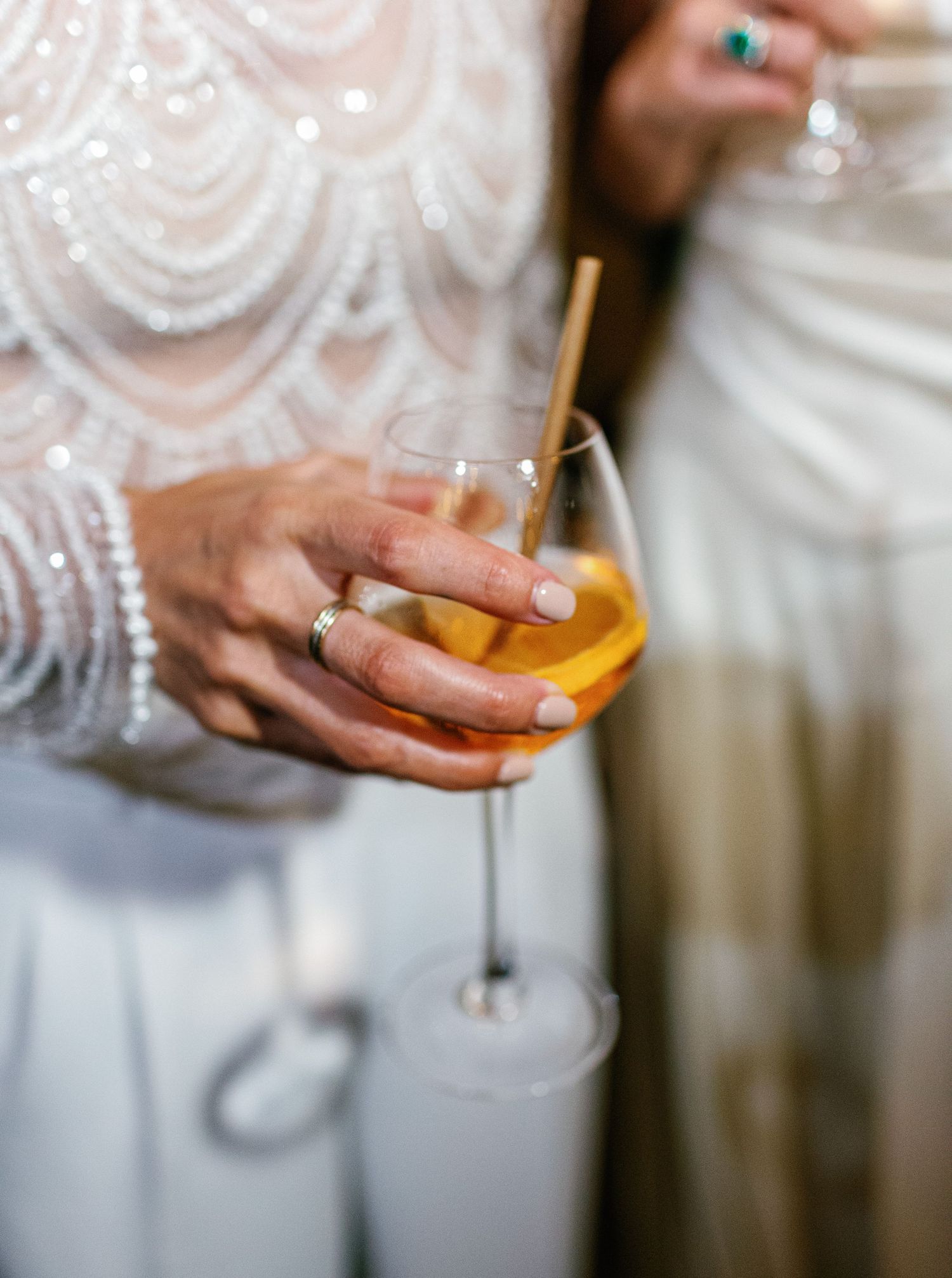 Close-up of hands holding a champagne glass filled with golden liquid while wearing a white sequined dress.