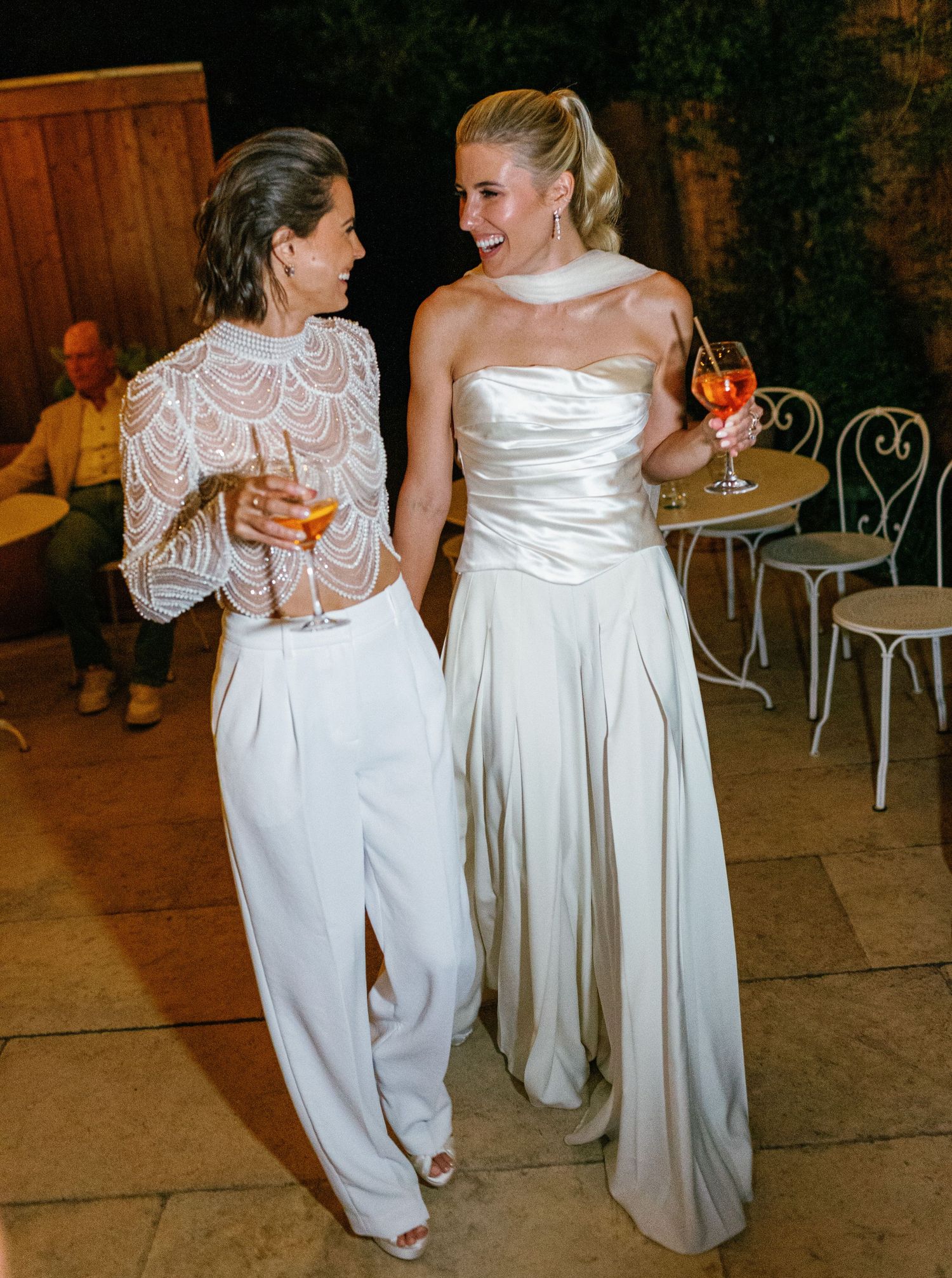 Two people in white formal attire sharing drinks and laughter at an elegant evening reception.