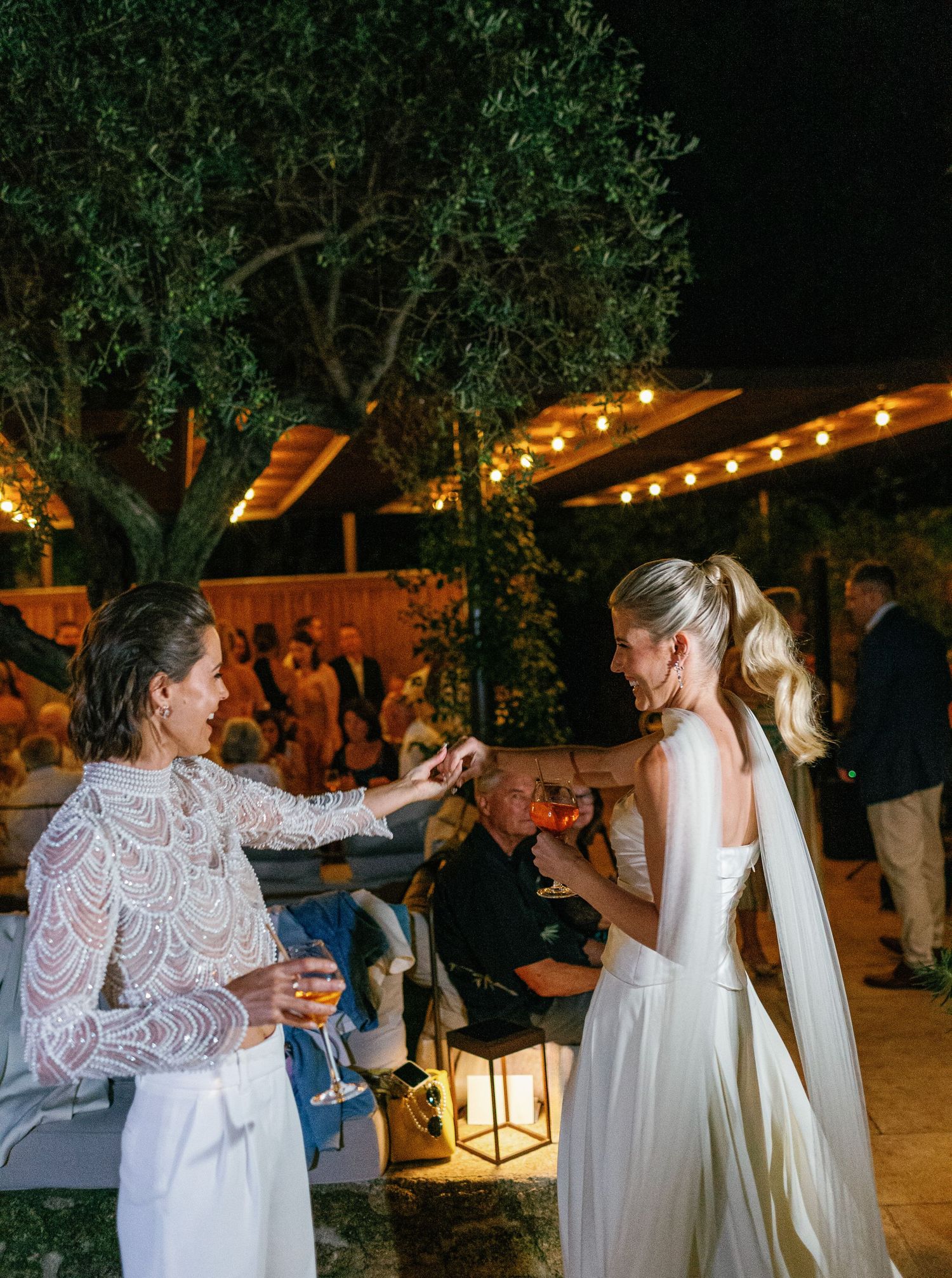 Wedding guests dance and celebrate on an outdoor patio decorated with string lights and trees at night.