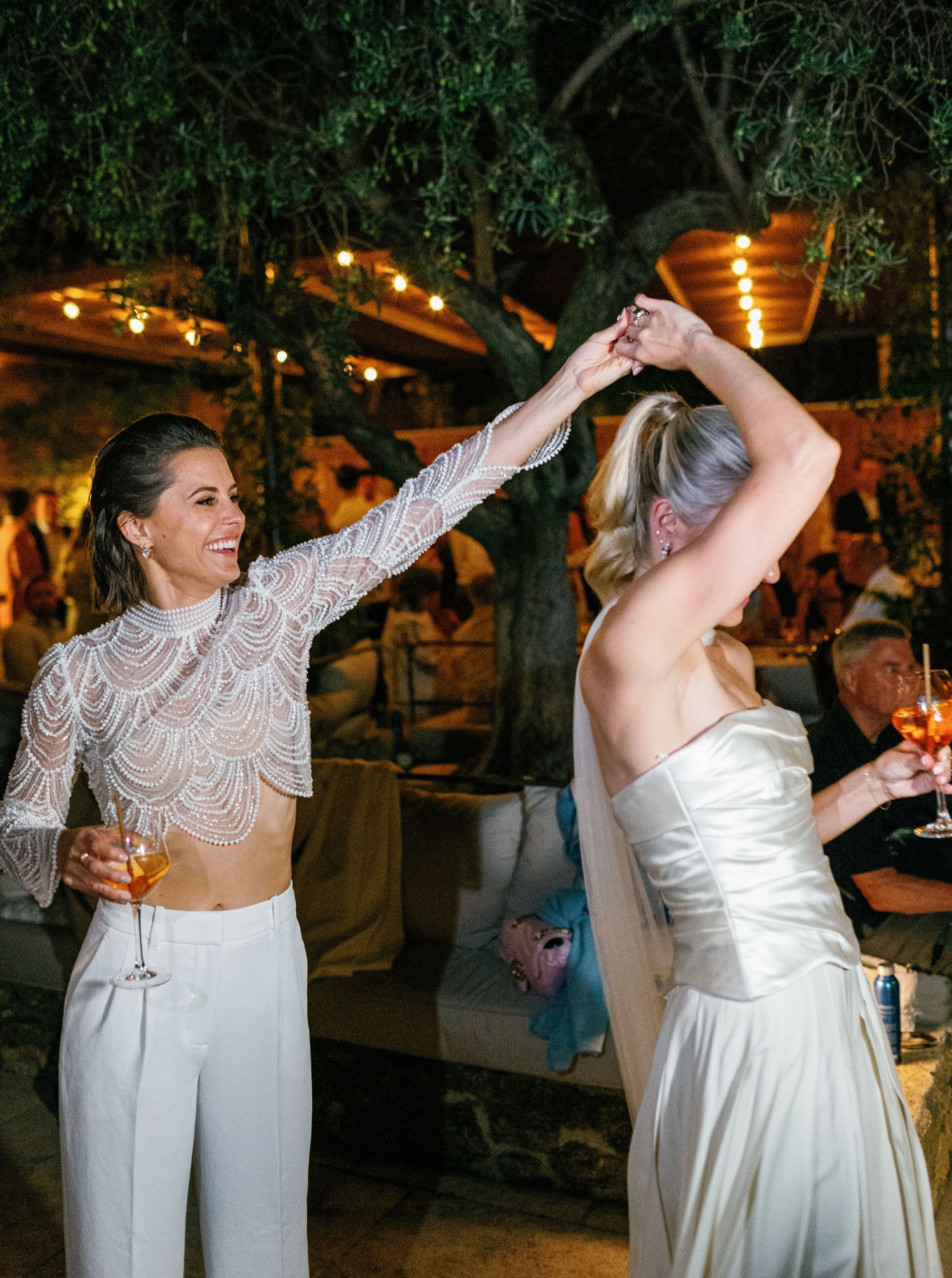 A couple in white attire dancing together at an evening outdoor reception with string lights overhead.