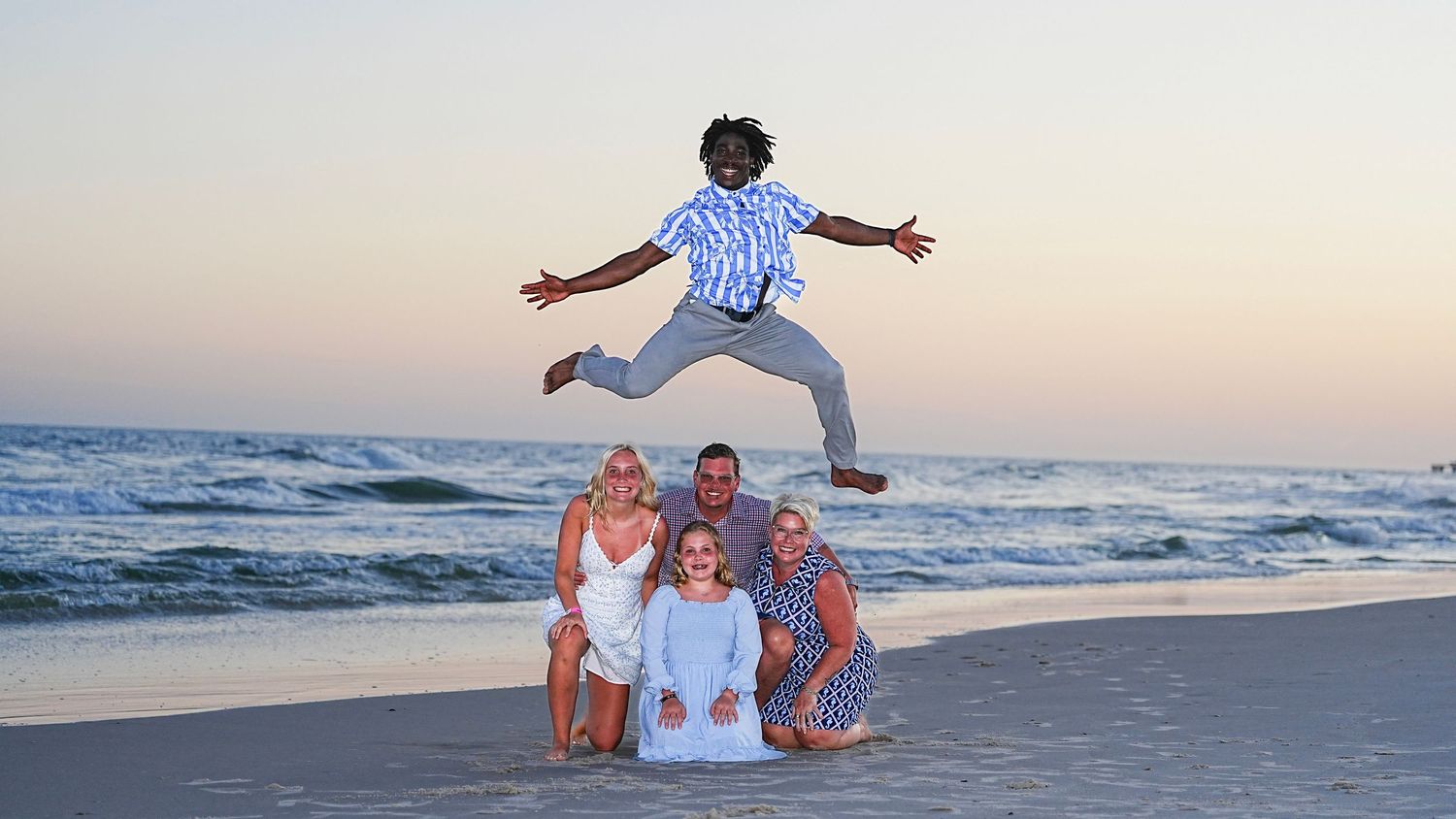 Family beach photo at sunset with child jumping in air above group posing on sandy shore near ocean waves.