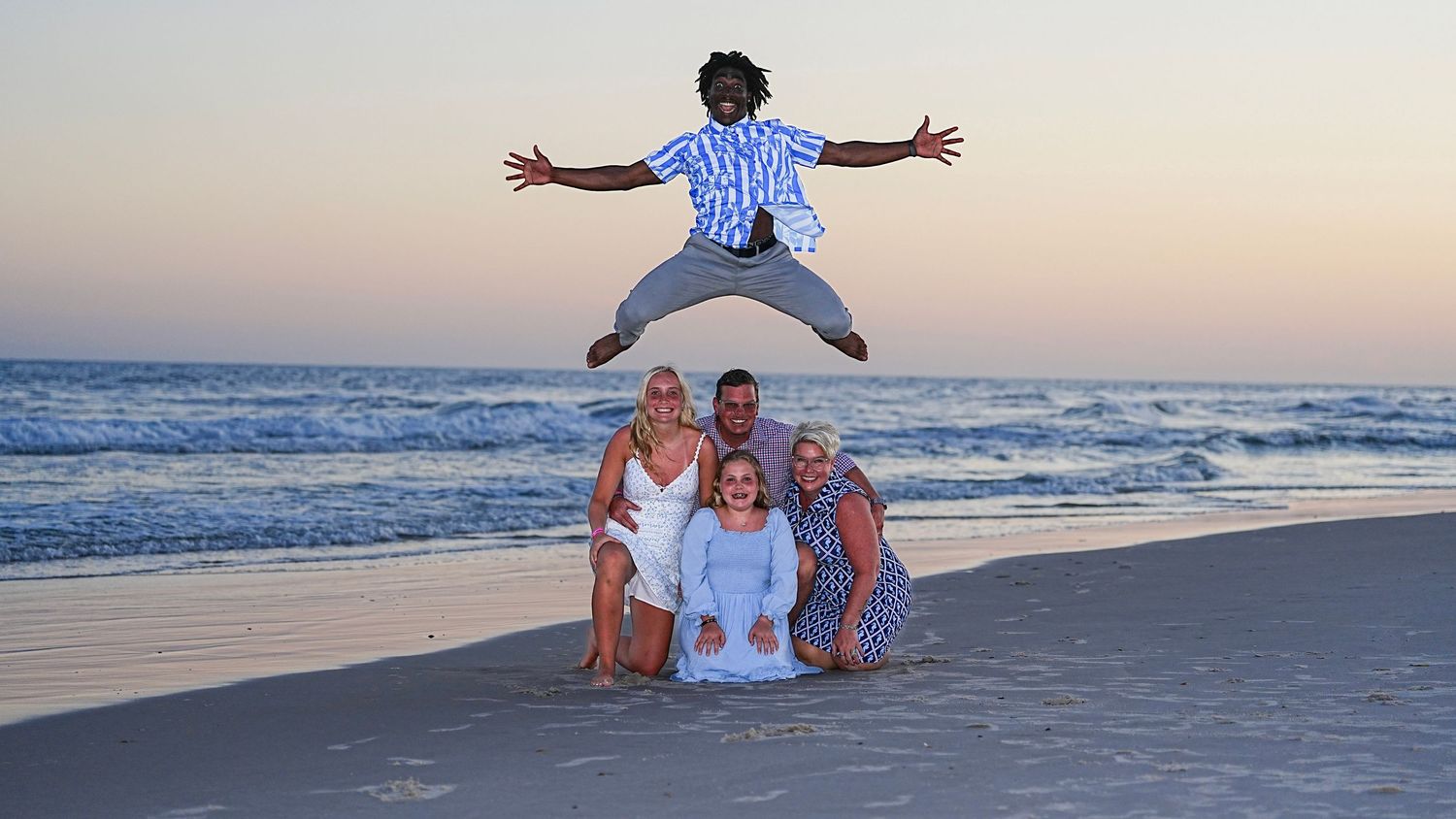 Family fun beach photo with person jumping in air above group posed on sandy shore at sunset.