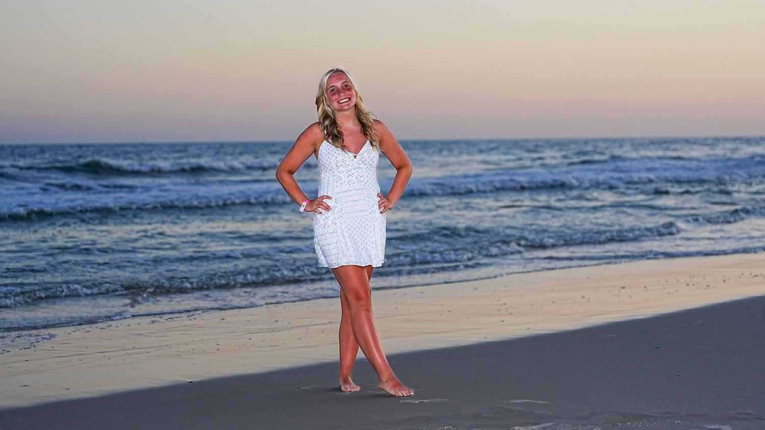 Woman in white dress posing on sandy beach at sunset with ocean waves in background.